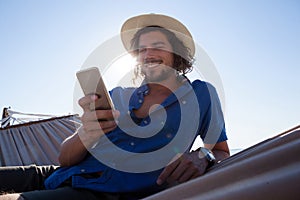 Smiling man using mobile phone while relaxing on hammock at beach