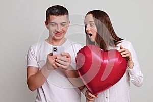 Smiling man using mobile phone scrolling online shocked woman looking at screen with surprised face sees shock content holding