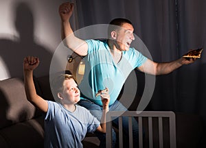 Smiling man is watching TV and cheering for football team with his son