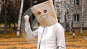 Smiling man wearing paper bag with happy emotion, man in mask standing outdoor and threatening his fist