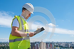 Smiling man in white helmet at construction site writes notes on clipboard