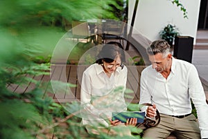 Smiling man and woman discussing business details and using tablet while sitting outdoors