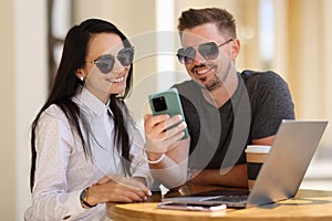 Smiling man and woman are sitting at table and looking into smartphone
