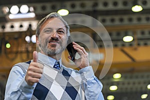 Smiling Manager Looking At Camera, Talking On Smartphone And Giving Thumb Up In A Factory