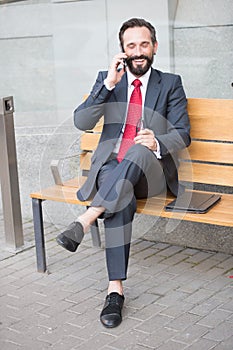 Smiling manager sitting on bench and phoning. Handsome smiled young businessman siting on bench with his laptop next to office