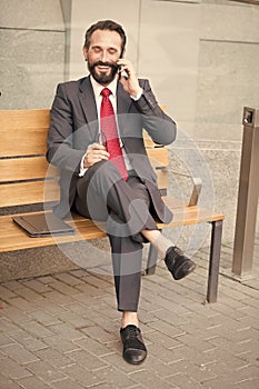 Smiling manager sitting on bench and phoning. Handsome smiled young businessman siting on bench with his laptop next to office