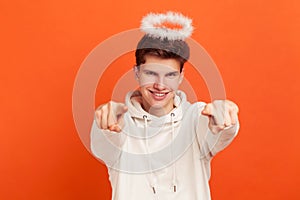 Smiling mannerly teenager in casual hoodie with halo overhead pointing fingers and looking at camera, sending rays of blessing and