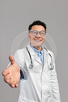 Smiling mature asian man doctor in white coat, glasses with stethoscope give his hand, greets patient in camera