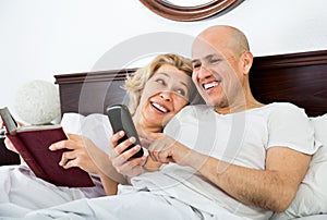 Smiling mature boyfriend and girlfriend lying in bed socialising
