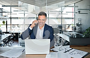 Smiling mature business man talking on cell phone using laptop in office.