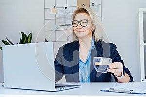 Smiling mature businesswoman working on laptop and drinking coffee at office, confident middle-aged experienced senior