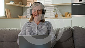 Smiling mature Caucasian woman grandmother businesswoman freelancer typing on laptop at home listening music on