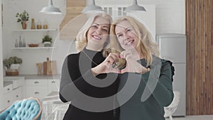 Smiling mature Caucasian women standing in luxurious kitchen and falding fingers into heart shape. Rich senior female