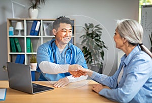 Smiling mature chinese man doctor shaking hands with elderly caucasian woman patient in clinic office