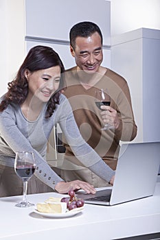 Smiling mature couple looking at laptop in the kitchen, drinking wine