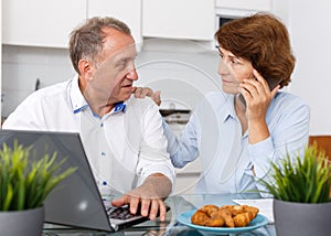 Smiling mature family couple with phone using laptop at kitchen table