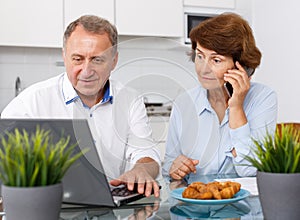 Smiling mature family couple with phone using laptop at kitchen table