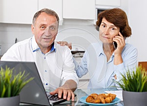 Smiling mature family couple with phone using laptop at kitchen table