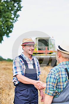 Smiling mature farmer shaking hands with senior farmer in field against harvester