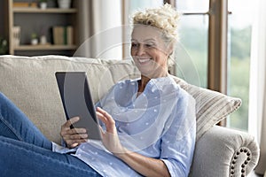 Smiling mature female resting on sofa with digital tablet