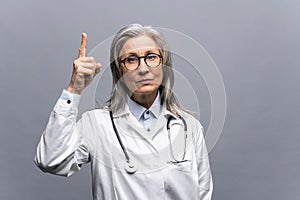 Smiling mature gray-haired female doctor in white medical gown, with stethoscope isolated on grey background points