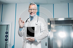 smiling mature male doctor with stethoscope over neck doing ok gesture and showing digital tablet with blank screen
