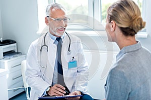 smiling mature male doctor writing in clipboard near female patient