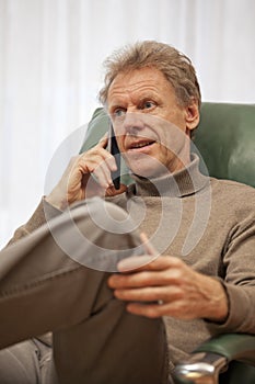 Smiling mature man talking on smartphone in a chair in a living room