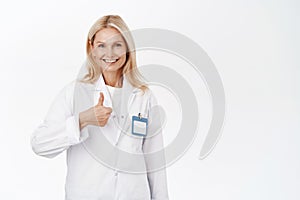 Smiling mature woman doctor, 50s years, shows thumbs up, recommends and promotes healthcare clinic, approves smth