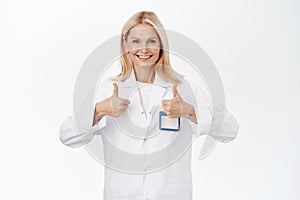 Smiling mature woman doctor, 50s years, shows thumbs up, recommends and promotes healthcare clinic, approves smth