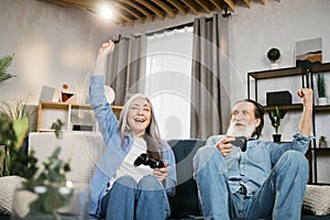 Smiling mature woman and her bearded husband sitting together on comfy couch playing video games