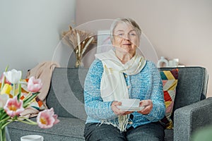 Smiling mature woman holding cup of tea, relaxing at home, positive senior female sitting on couch in modern living room