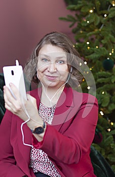 Smiling mature woman is looking into camera while holding her smart phone in a hand near Christmas spruce tree.