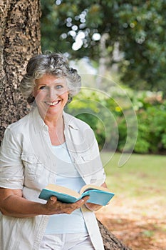 Smiling mature woman reading book leaning on tree trunk
