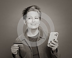Smiling matured woman in casual clothes holding cellphone and bank card in hands isolated on grey background in balck