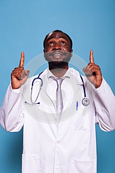 Smiling medical doctor pointing both index fingers up and wearing lab coat and stethoscope