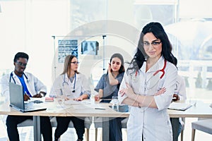 Smiling medical doctor woman with stethoscope standing in front of medic team at hospital. Attractive young female