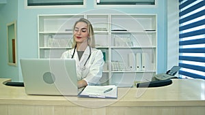 Smiling medical nurse working on laptop and making notes at reception desk