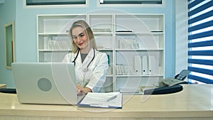 Smiling medical nurse working on laptop and making notes at reception desk