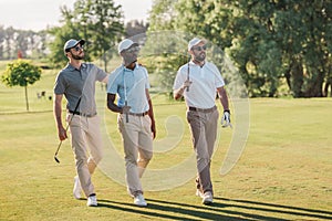 Smiling men in caps and sunglasses holding golf clubs and walking on lawn