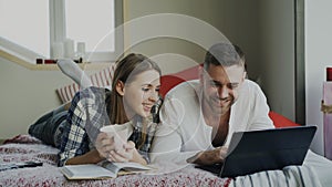 Cheerful man and woman talking and lying in bed reading book and using laptop computer in the morning