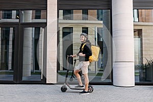 Smiling messneger man standing outdoors with electric push scooter. Delivery man with termal backpack in the city photo