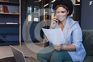 Smiling mid adult biracial businesswoman talking on smart phone while reading document in office