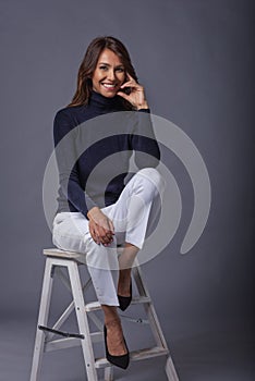 Smiling mid aged brunette woman sitting on a stool at isolated gray background