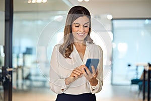 Smiling mid aged business woman executive using smartphone standing in office.
