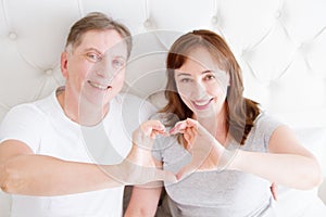 Smiling middle age couple showing heart sign by arms in bedroom. Love and family life relationship concept. Selective focus