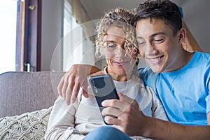Smiling middle-aged 40s mother rest with grown-up son using smartphone together, happy young man enjoy family weekend with mom