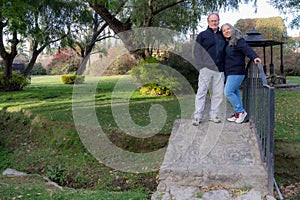 Smiling middle aged adult couple standing on small bridge with green foliage trees in background