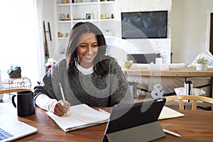 Smiling middle aged African American  woman sitting at the table in her dining room looking at tablet computer and making notes, s