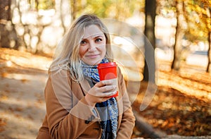 Smiling middle aged blonde woman sitting in city park on sunny autumn day with cup of coffee to go